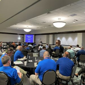 Atendees at the 2021 PIPM Summit are seated around circular tables, listening to the keynote speaker in the front of the room.